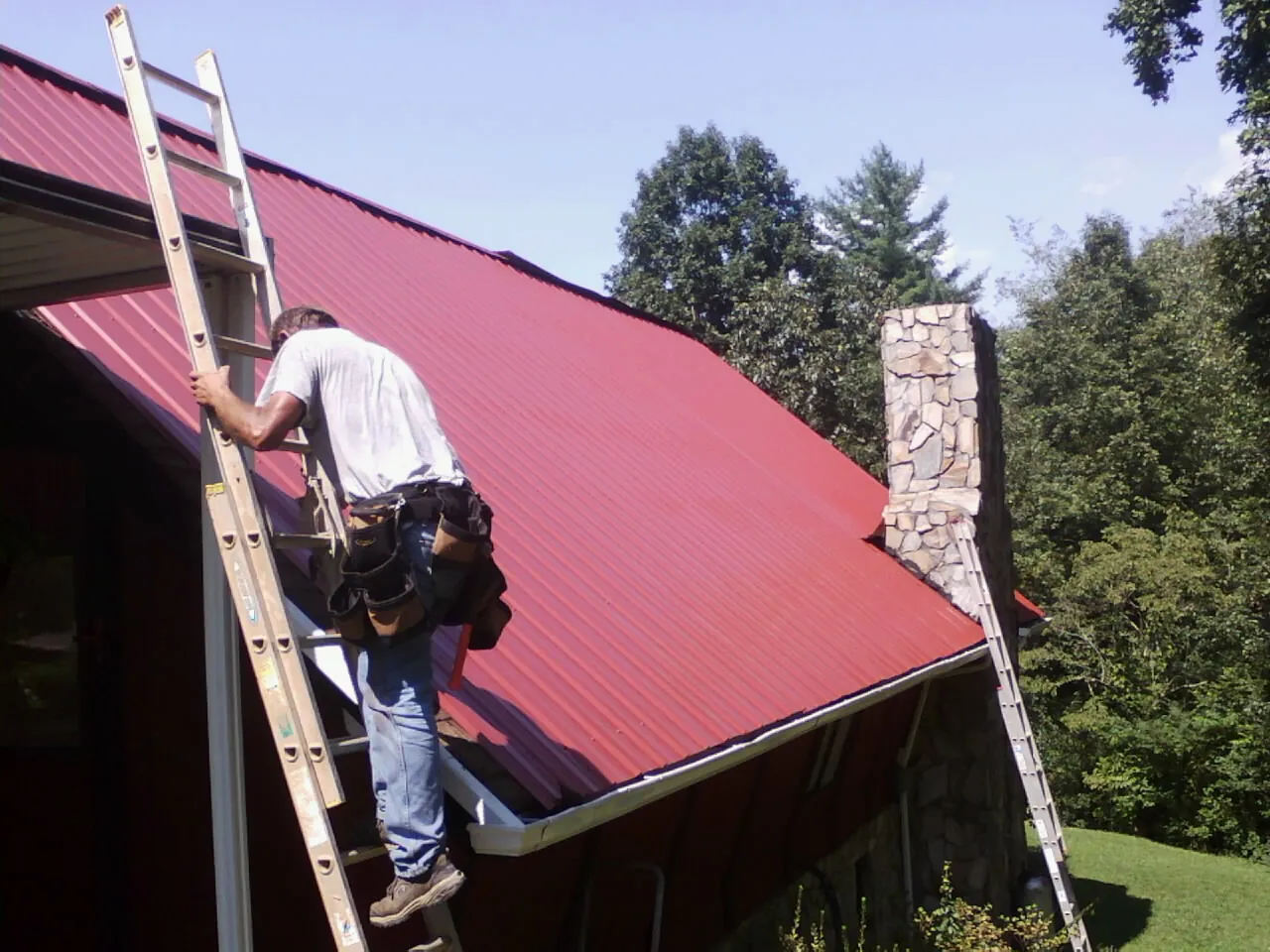Roofer on a ladder beside a red metal roof and stone chimney — Soffit & Fascia Repair by Roofing Brothers in Stamford